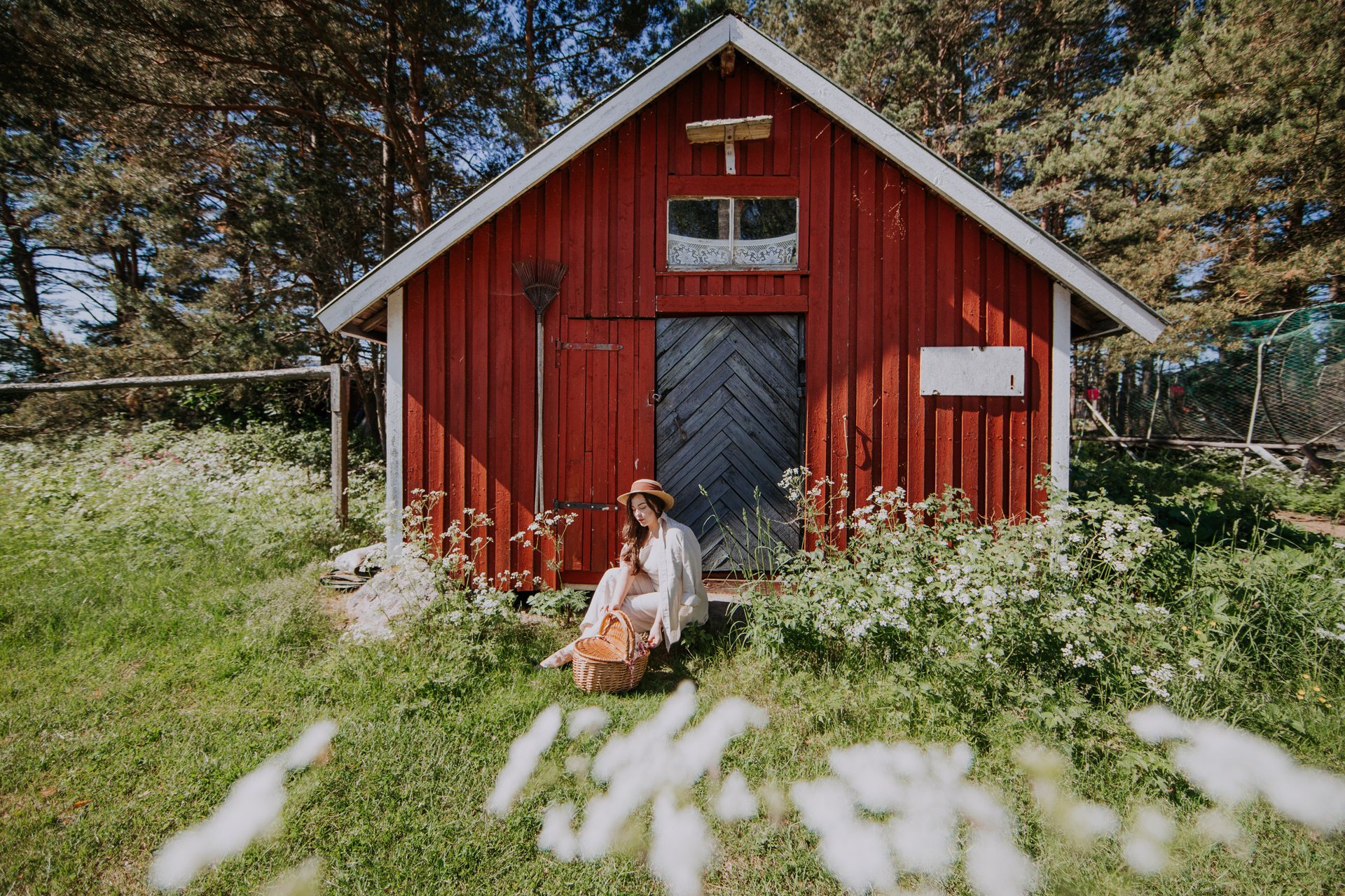 Nainen istuu punaisen lautamökin edustalla kesäisessä maisemassa Kaunissaaressa. Mökin seinä on maalattu kirkkaan punaiseksi, ja siinä on valkoiset nurkkalaudat. Naisen vieressä on punottu kori ja hänellä on päässään leveälierinen hattu. Mökin ympärillä kasvaa vehreää ruohoa ja valkoisia kukkia, jotka tuovat esille paikan luonnonläheisyyden. Taustalla näkyy metsää, ja aurinko paistaa kirkkaalta siniseltä taivaalta, luoden lämpimän ja kutsuvan tunnelman.