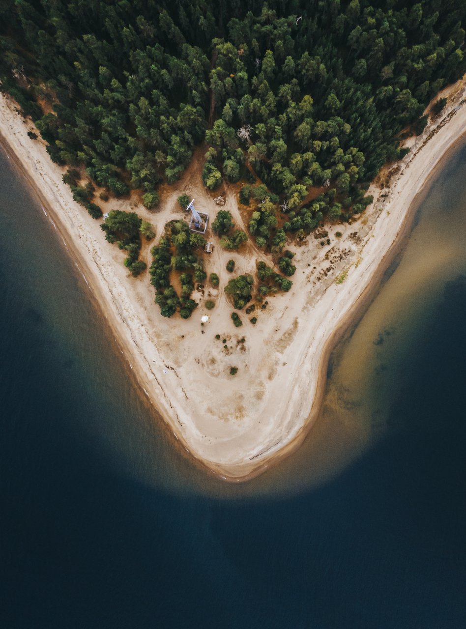 Ilmakuva Kaunissaaresta, joka on otettu dronella. Kuvassa näkyy saaren metsäinen kärki, jota ympäröi hiekkaranta ja kirkas vesi. Metsän keskellä on näkyvissä polkuja ja pieni rakennus. Vesi on syvän sinistä ja rannikko matalaa vaaleaa hiekkaa.