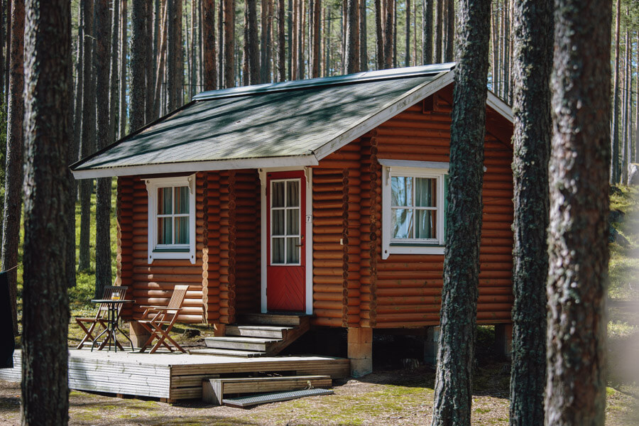 Valokuva pienestä punaisesta hirsimökistä, joka sijaitsee metsän keskellä korkeiden mäntyjen ympäröimänä. Mökissä on valkoiset ikkunanpuitteet ja punainen ovi. Mökin edessä on pieni terassi, jossa on kaksi tuolia ja pöytä.