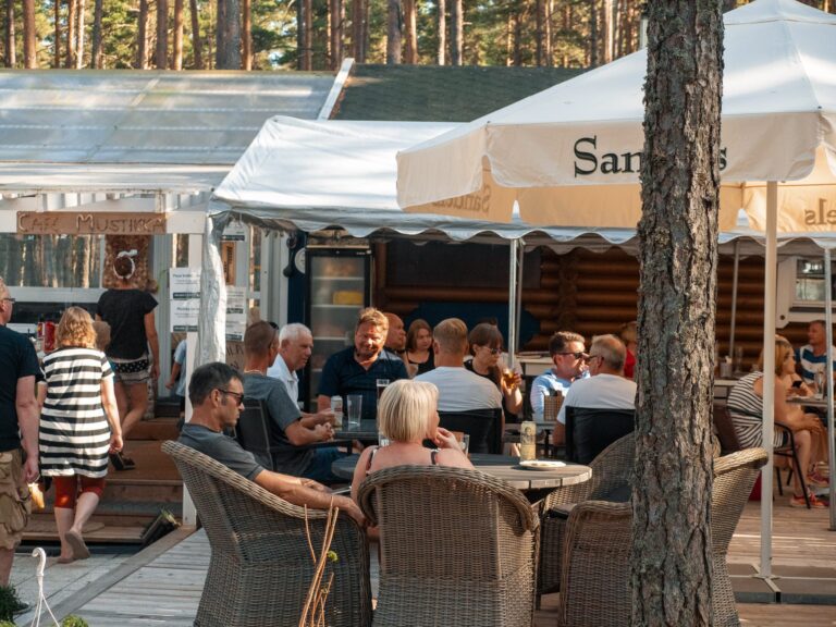 Valokuva ulkoilmakahvilasta, jossa ihmiset istuvat ja nauttivat juomia ja ruokaa. Useat pöydät ovat varjoisien aurinkovarjojen alla. Taustalla näkyy kyltti 'Cafe Mustikka' ja useita ihmisiä seisomassa ja kävelemässä. Ympärillä on mäntymetsä.