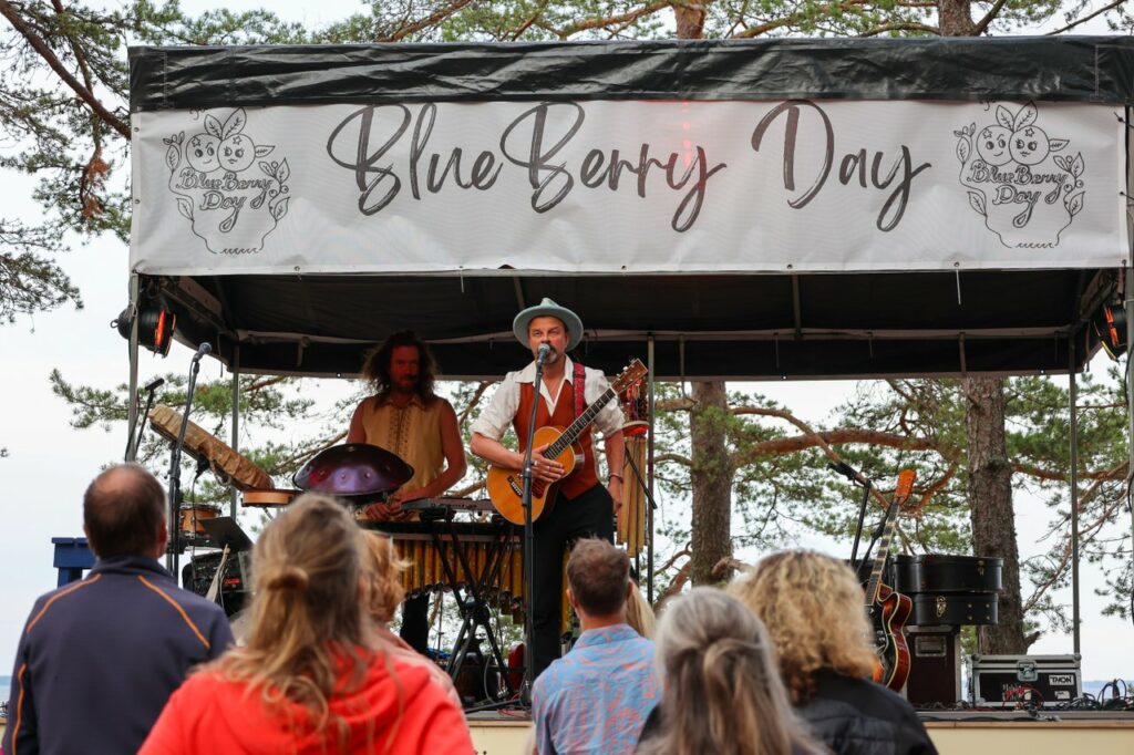 Valokuva BlueBerry Day -festivaalilta 2023. Lavalla esiintyy muusikko, joka soittaa kitaraa ja laulaa, taustallaan toinen muusikko, joka soittaa lyömäsoittimia. Lavalla on suuri kyltti, jossa lukee 'BlueBerry Day'. Etualalla näkyy festivaaliyleisöä, joka katsoo esiintyjiä. Taustalla näkyy puita.